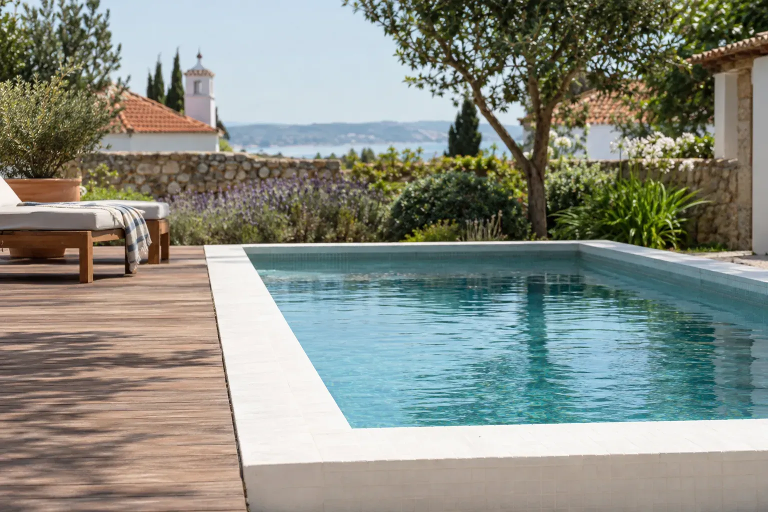 Piscina retangular com remate branco, junto a um deque de madeira e uma oliveira, com vista para o mar.